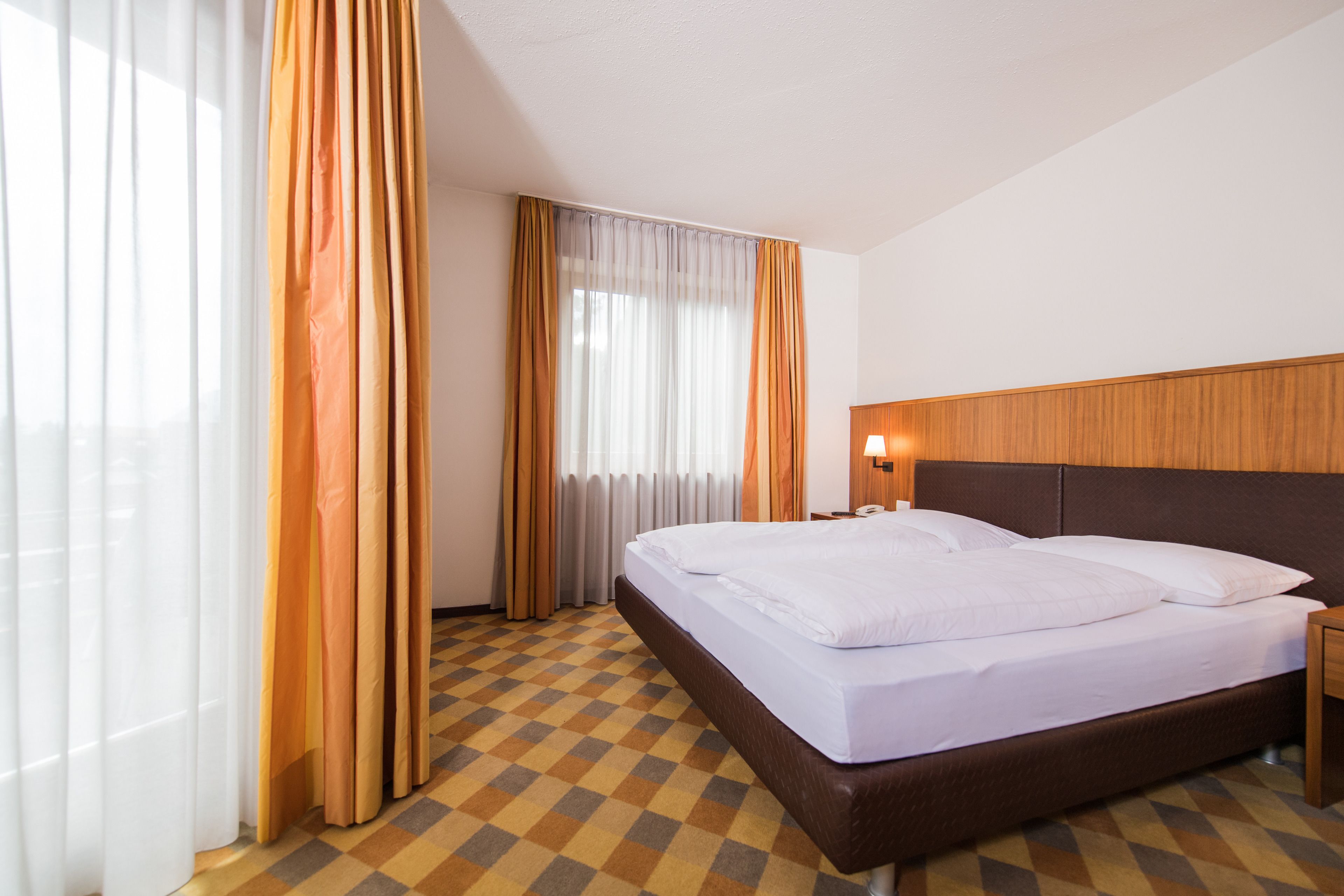 A hotel room with a large bed, a wooden headboard, a nightstand, and a window with curtains.