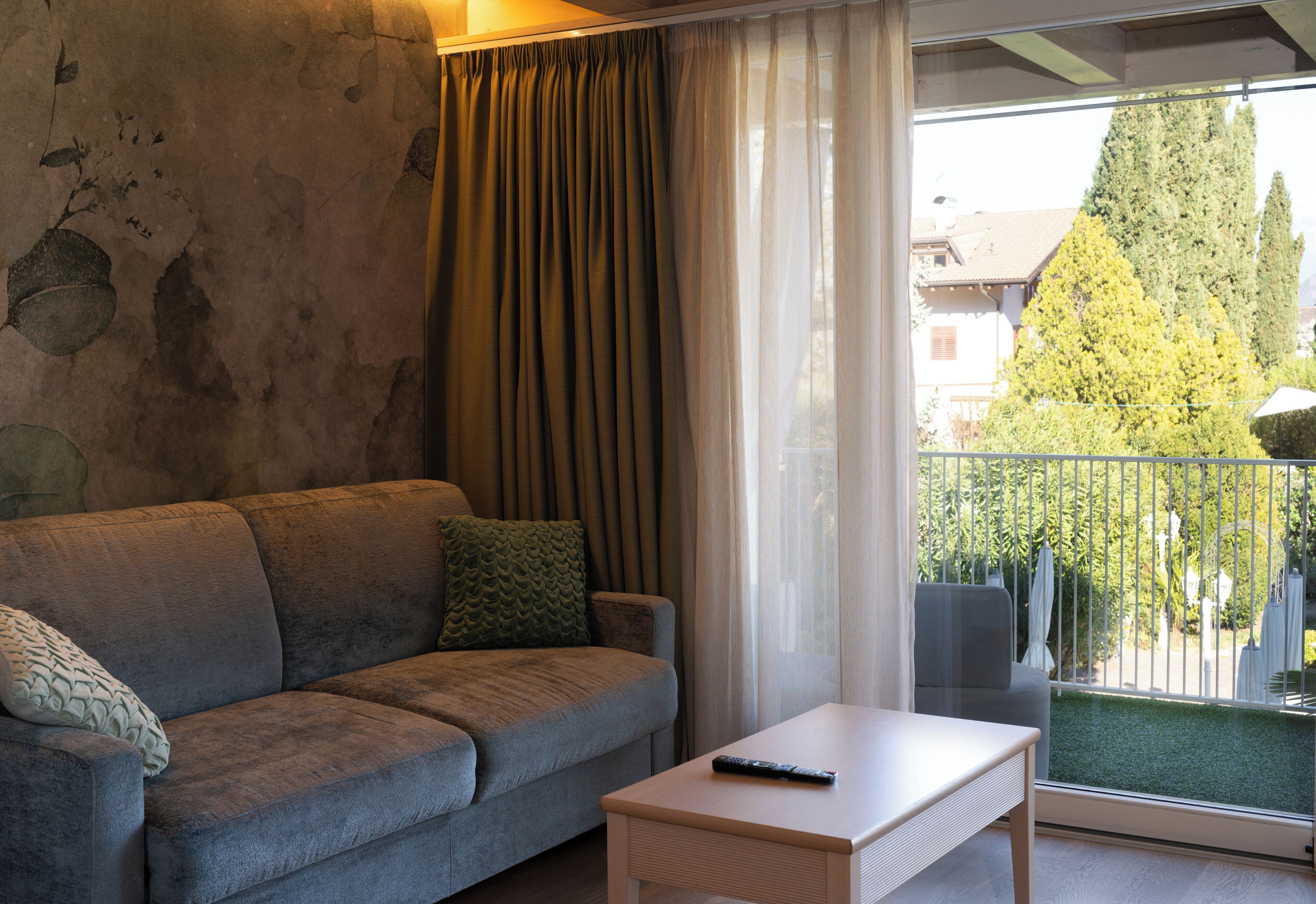 Un salón con sofá gris, una mesita blanca y un balcón con vistas a árboles y edificios.