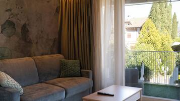 Un salón con sofá gris, una mesita blanca y un balcón con vistas a árboles y edificios.