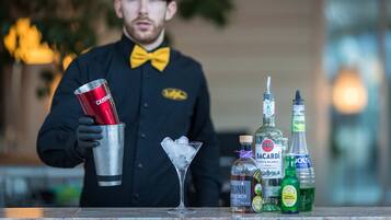 Un barman sosteniendo un agitador de cócteles y una copa de martini, con botellas de licor y una botella de agua sobre la barra.