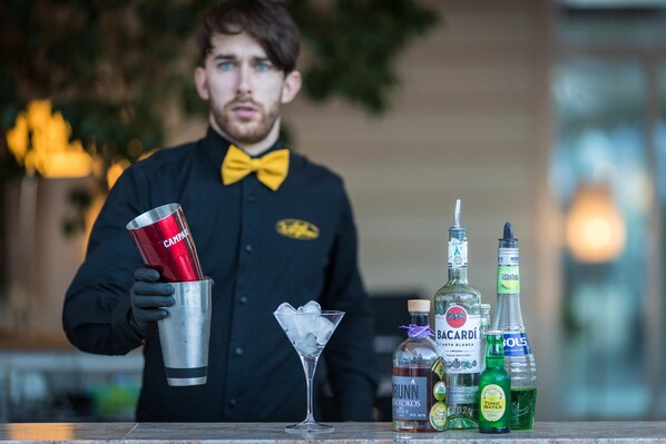 A bartender holding a cocktail shaker and a martini glass, with bottles of alcohol and a water bottle on the counter.