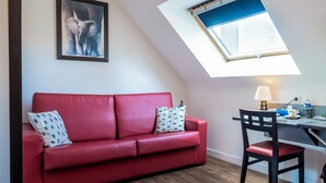 A red sofa with patterned pillows, a wooden desk with a lamp, and a skylight above. - Auberge du Cheval Blanc (Baud)