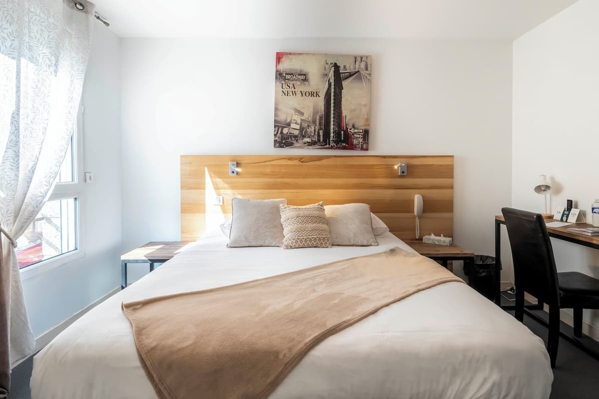 A hotel room with a bed, a desk, a chair, a lamp, and a wall-mounted artwork.