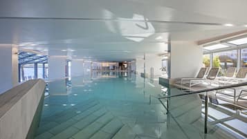 A modern indoor swimming pool with clear glass walls, a geometric floor pattern, and a seating area with lounge chairs.