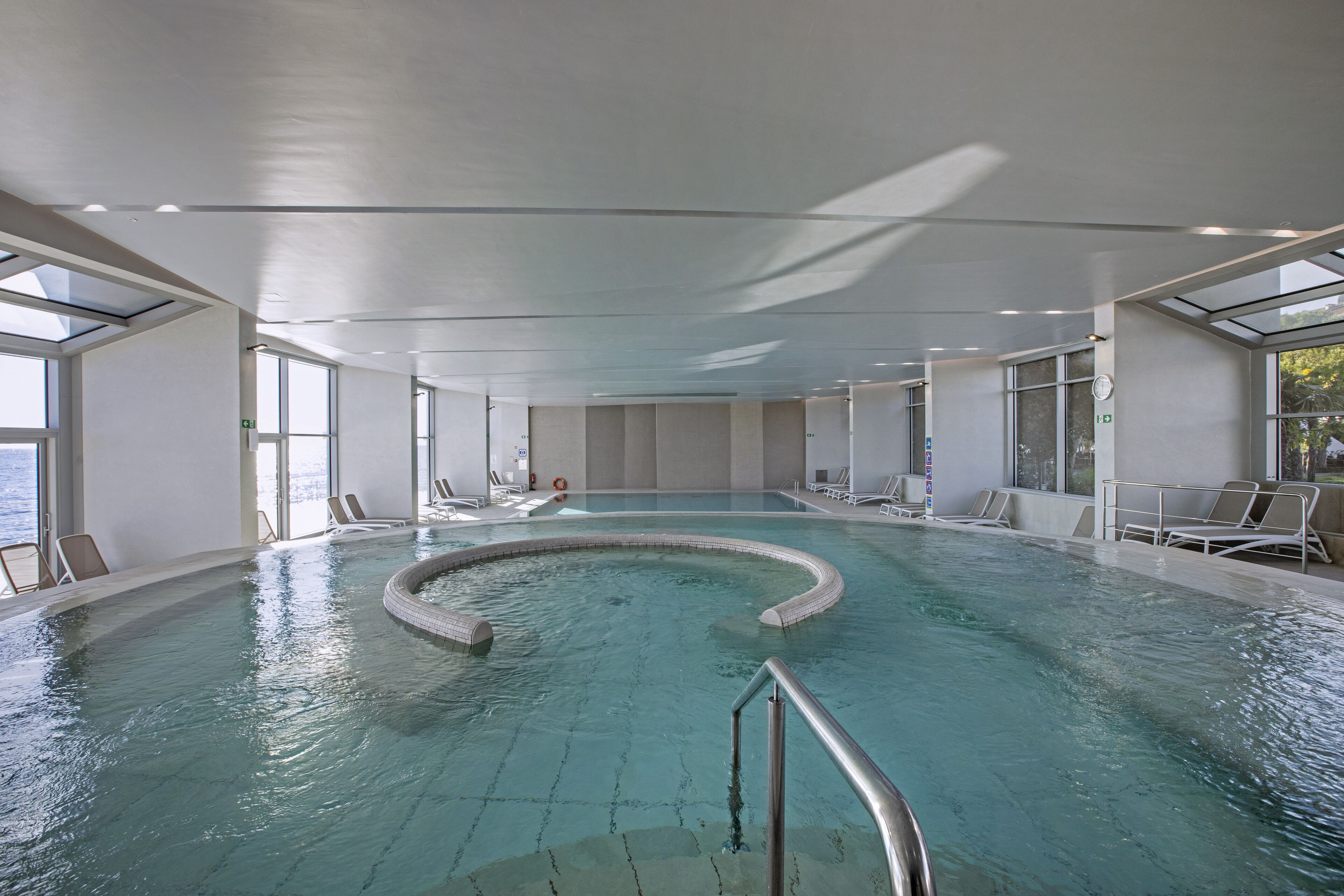 Une grande piscine intérieure à la forme incurvée, entourée de chaises longues et dotée d’un plafond en verre.