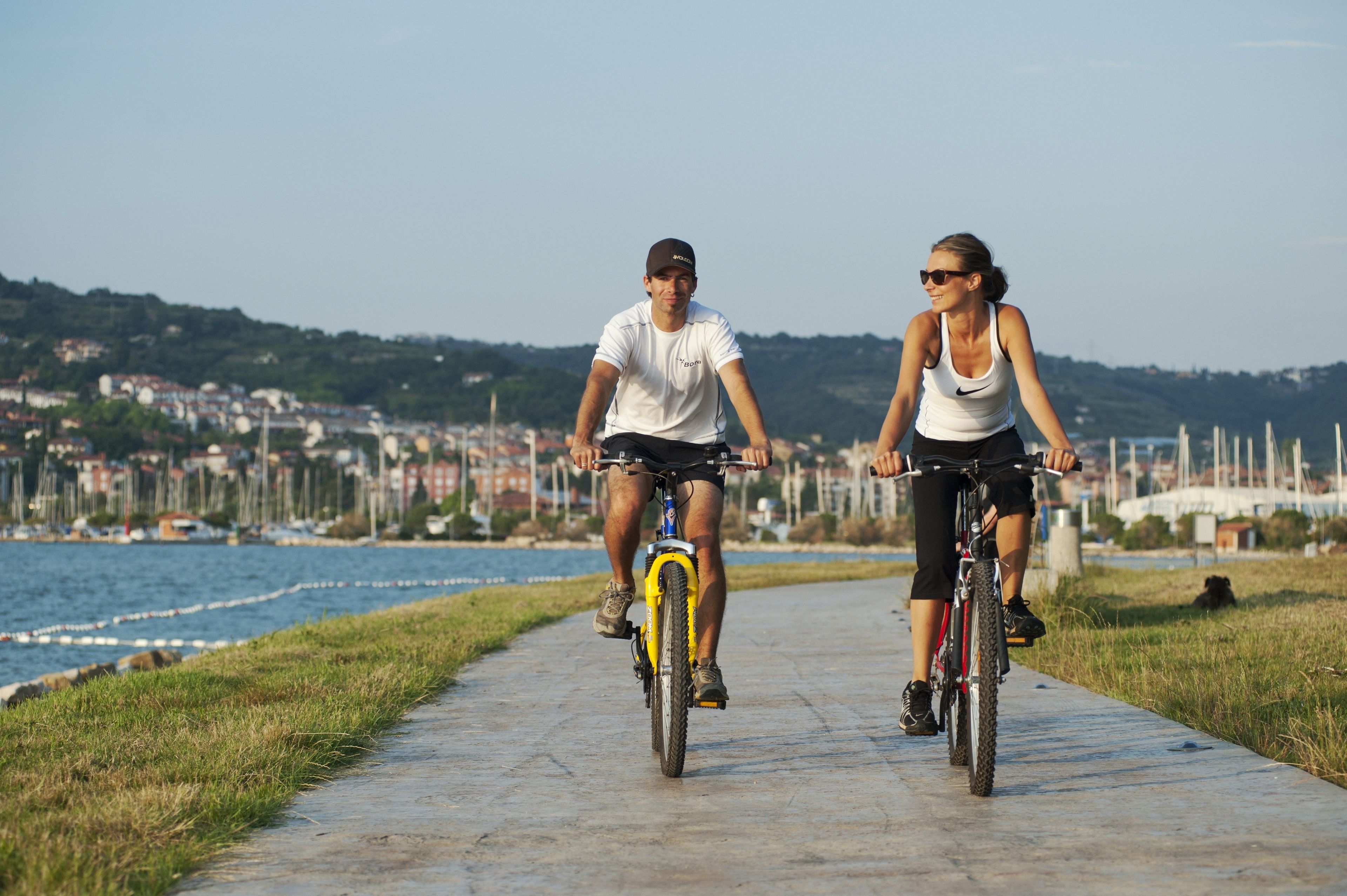 Two people riding bicycles on a concrete path by a waterfront with buildings in the background.