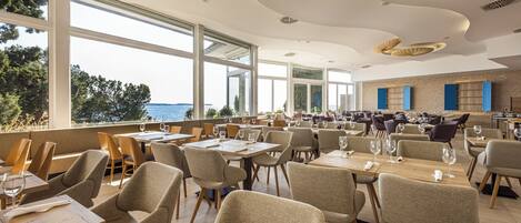 A spacious restaurant with large windows offering a view of the sea. Tables are neatly arranged with chairs, and the ceiling features unique geometric patterns.