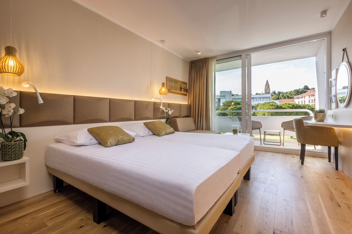 A modern hotel room with a large bed, a desk, and a balcony with a view of the city.