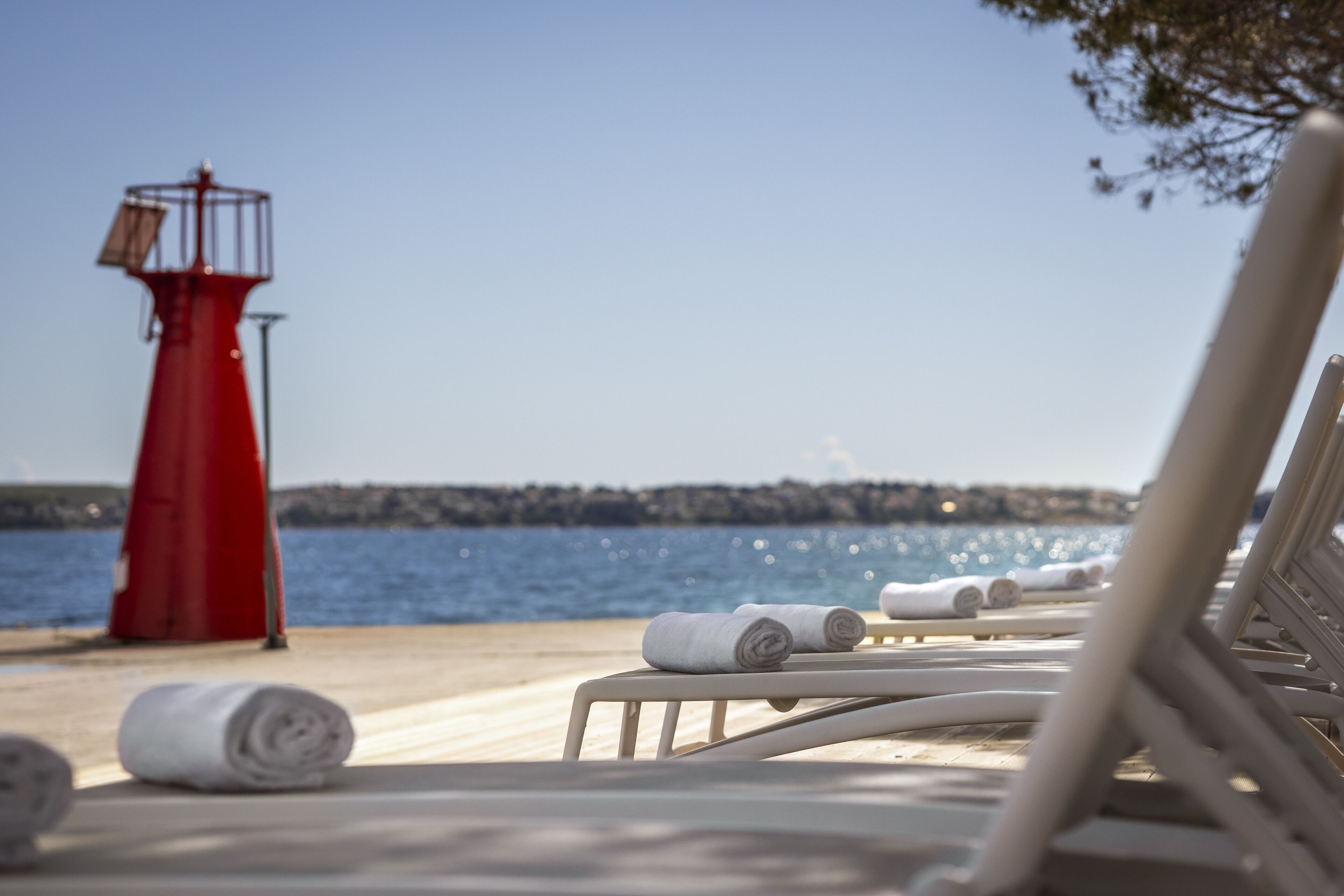 A beachside lounge with sun loungers and towels, a red lighthouse in the background, and a clear view of the sea.