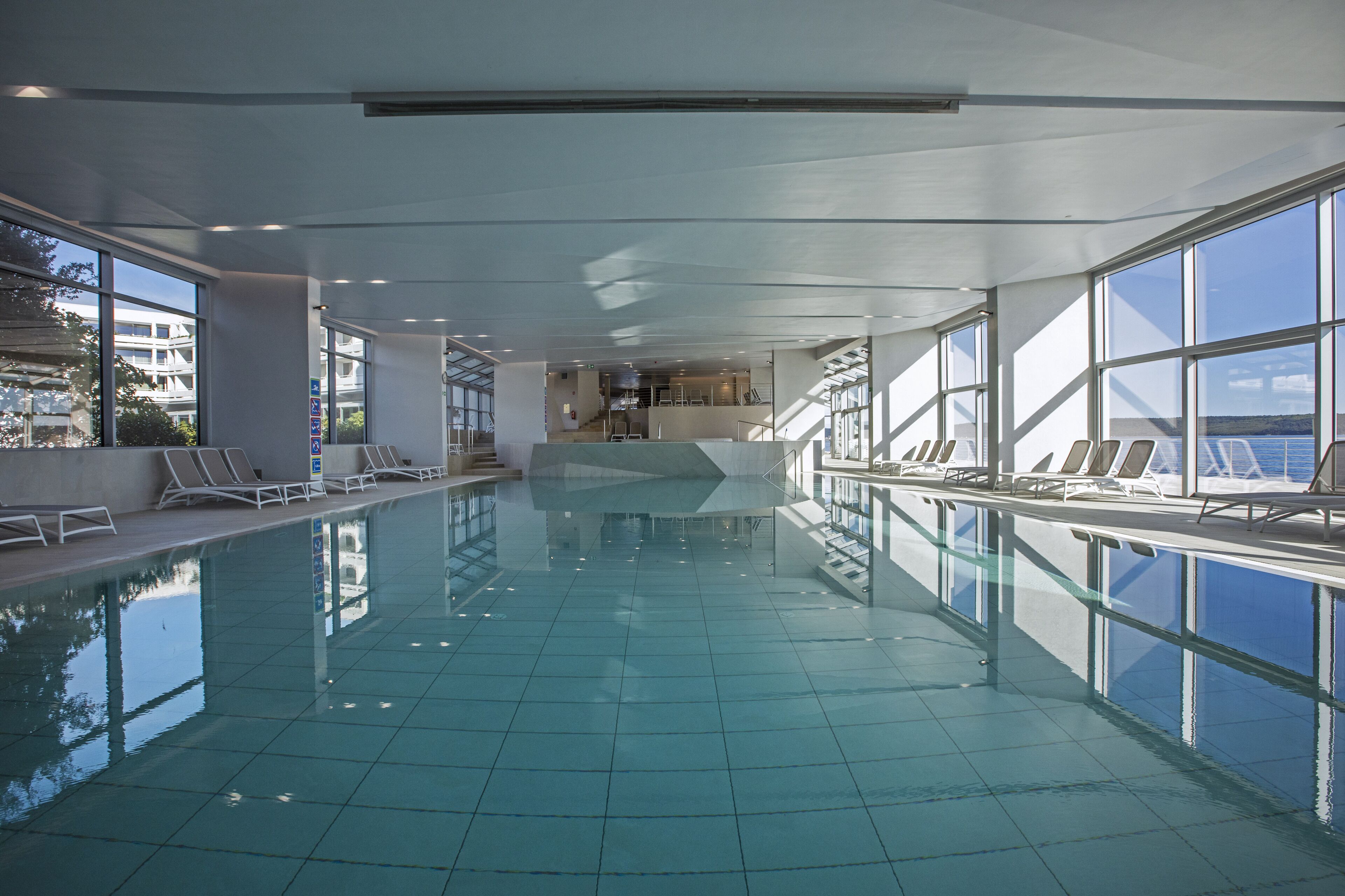 An indoor swimming pool with clear glass walls, a tiled floor, and sunlit interior.