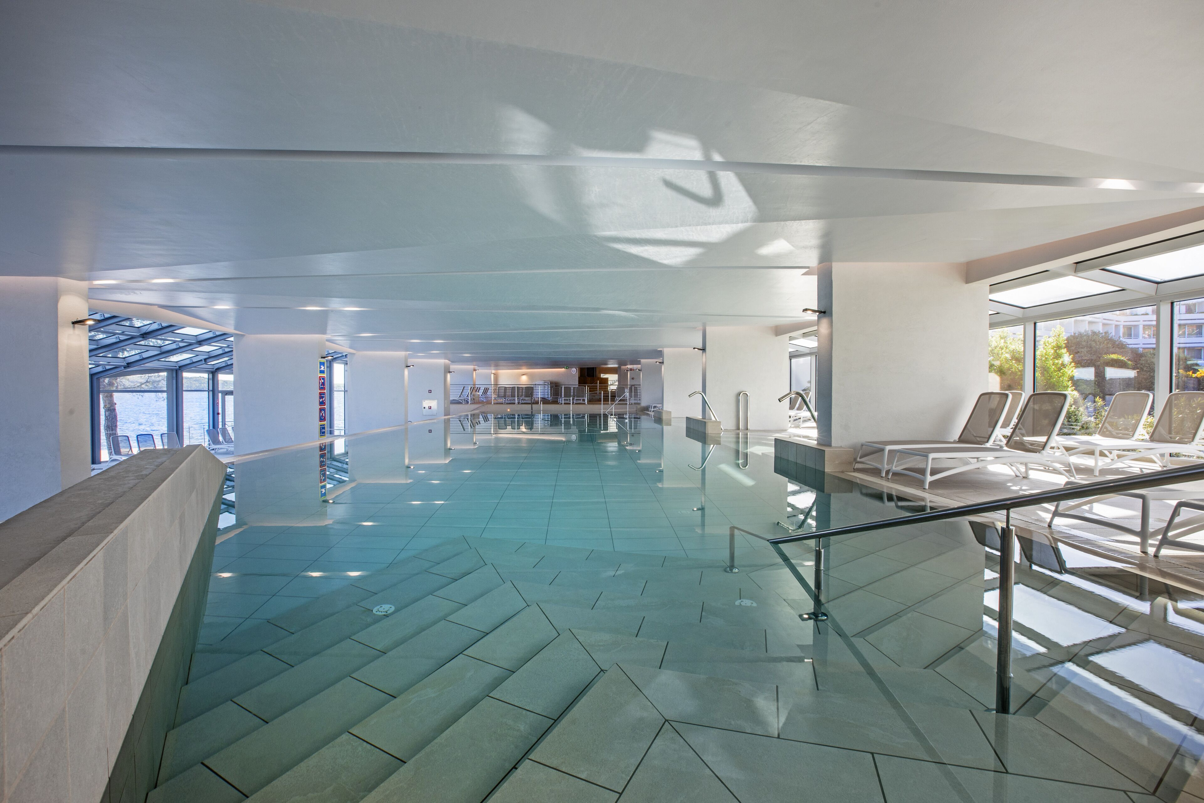 Ein moderner, überdachter Swimmingpool mit klaren Glaswänden, einem geometrischen Bodenmuster und einer Liegebereich mit Liegestühlen.