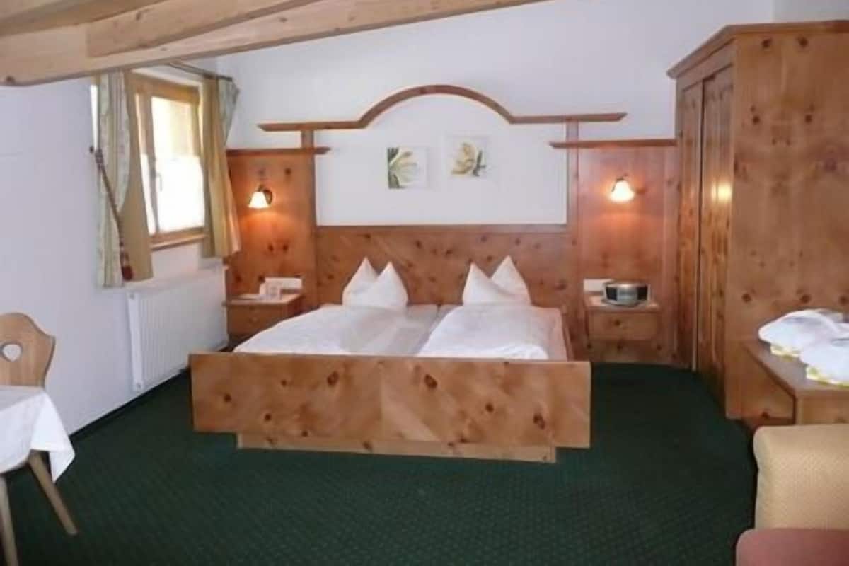 Uma cama de madeira com cabeceira, duas mesas de cabeceira, uma cadeira e uma janela com cortinas.
