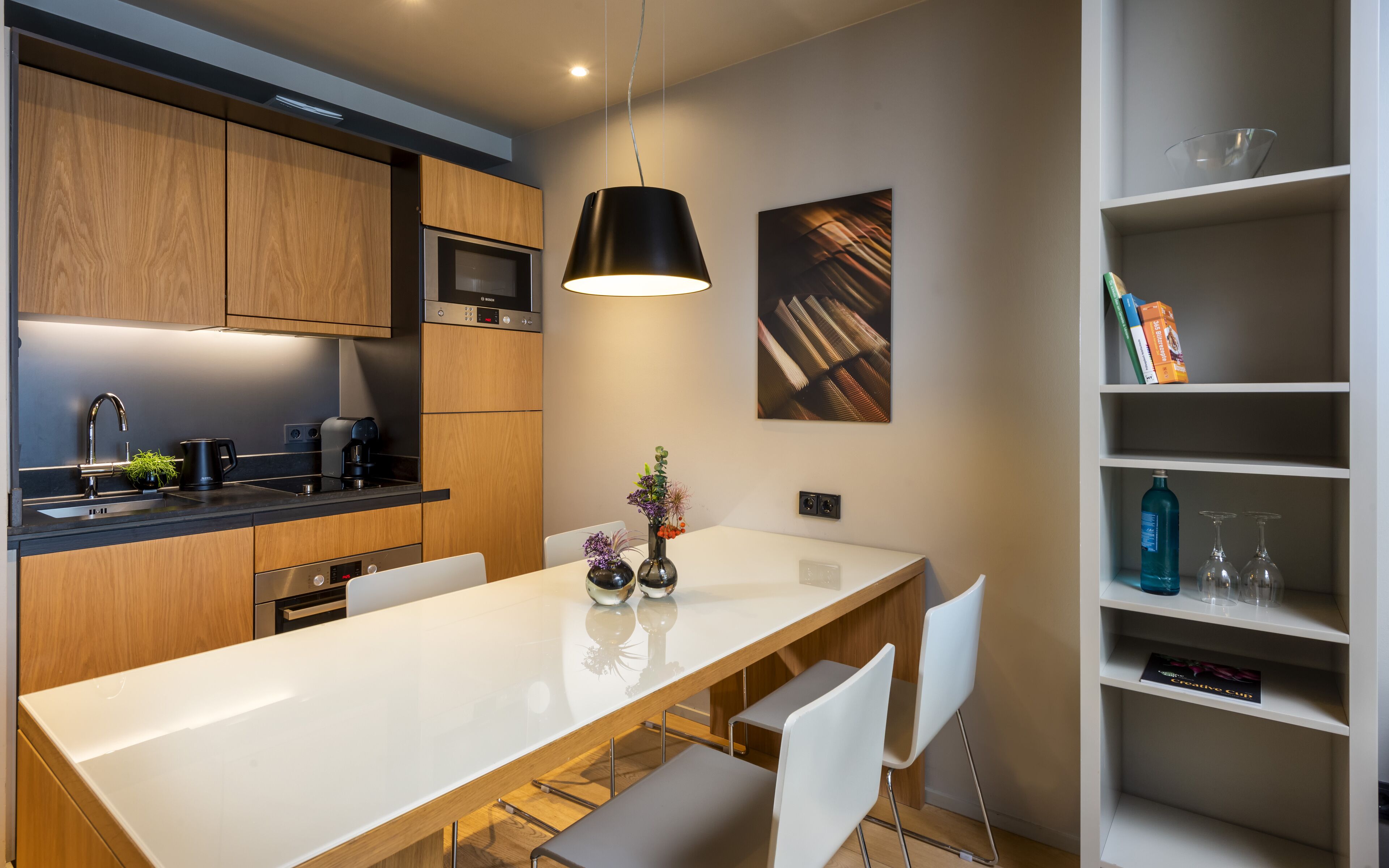 A modern kitchen with wooden cabinets, a Bosch microwave, a black hanging light, and a white dining table with chairs.