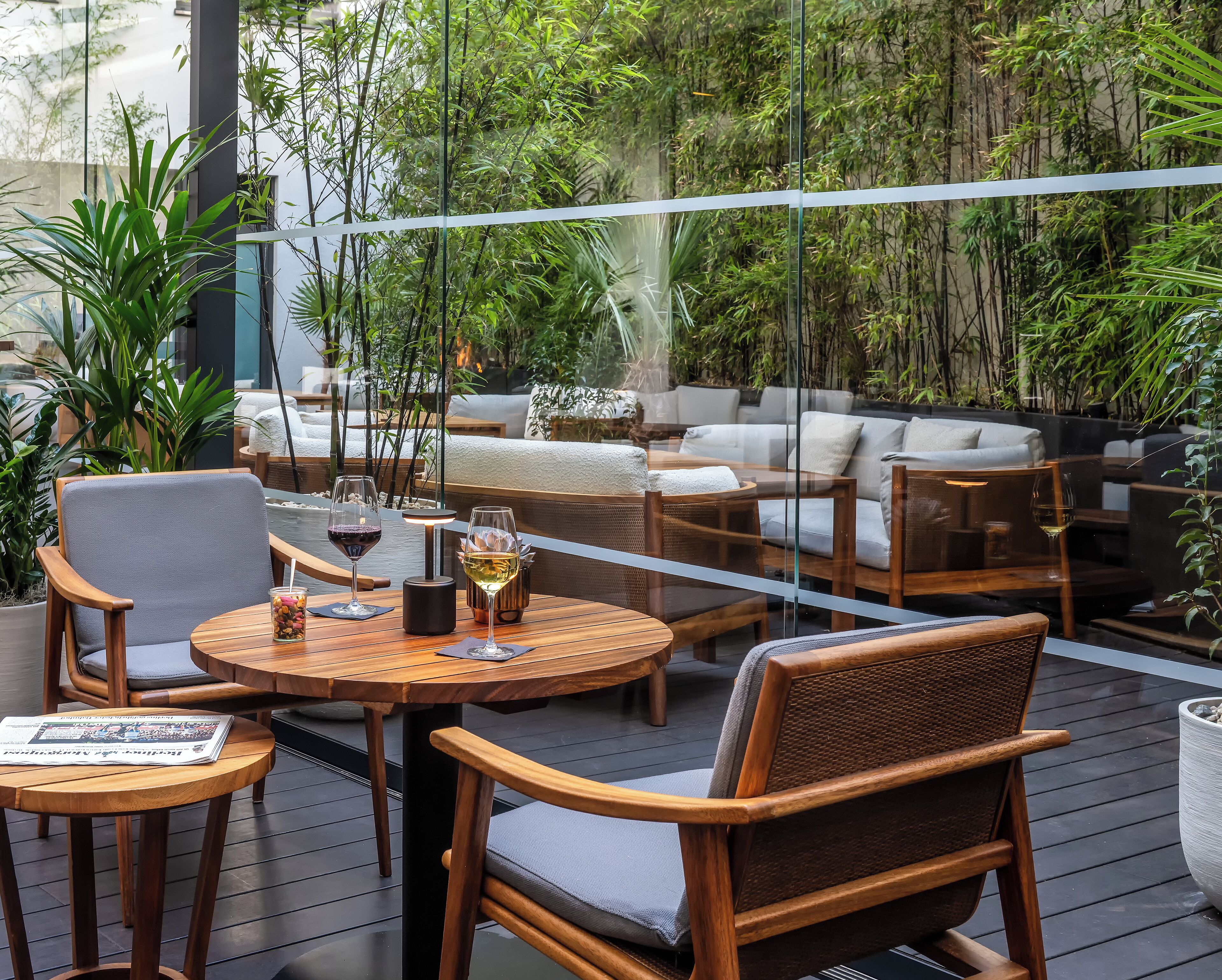 An outdoor seating area with wooden furniture and glass tables, surrounded by lush greenery.