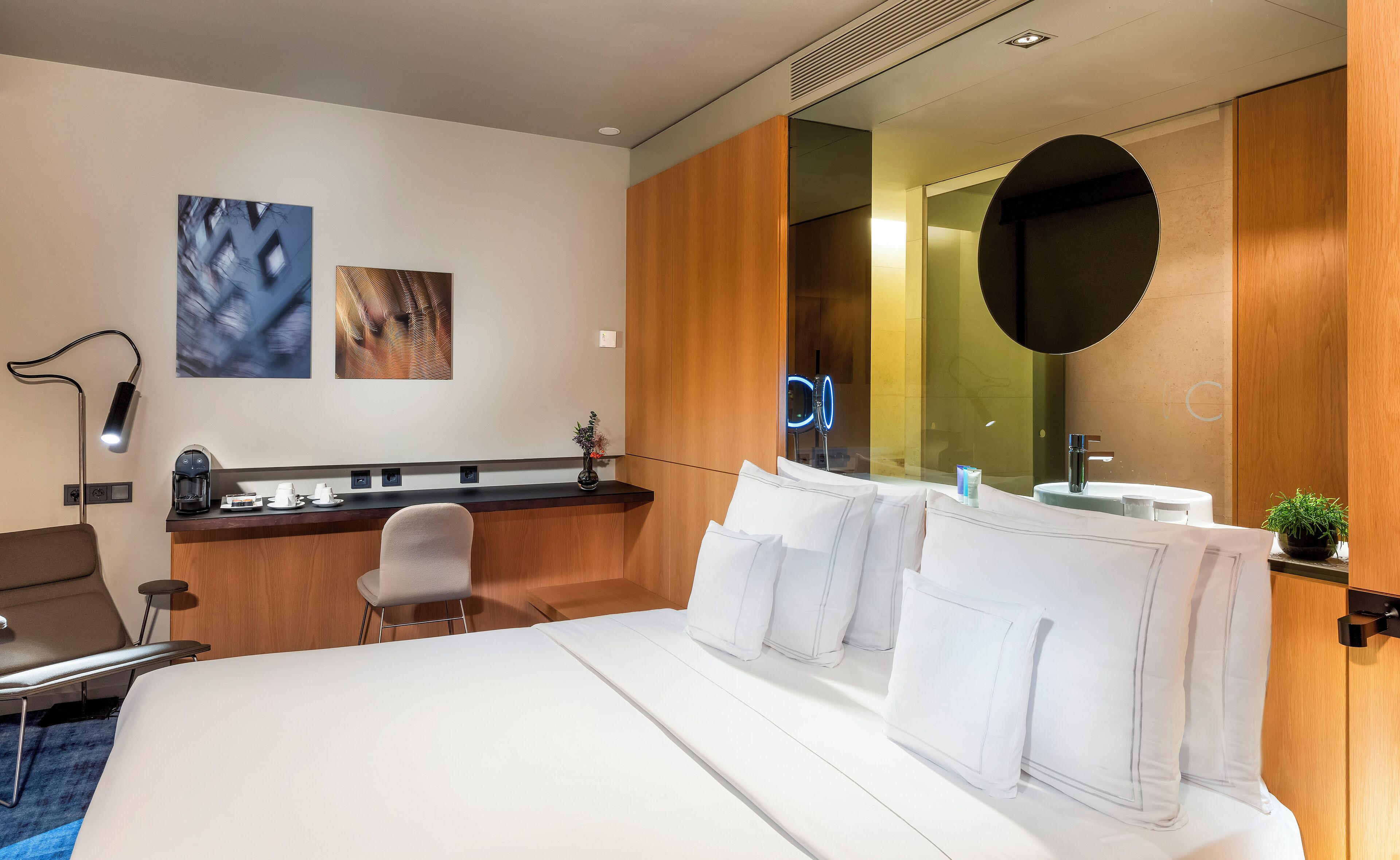 Habitación de hotel con cama, escritorio, silla, lámpara de escritorio, cafetera, espejo y una planta pequeña.