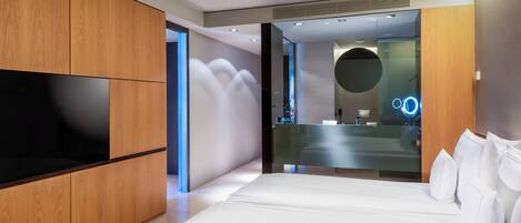 A modern bedroom with a large bed, wooden wardrobe, and a glass partition separating the room from a bathroom with a circular mirror and neon lights.