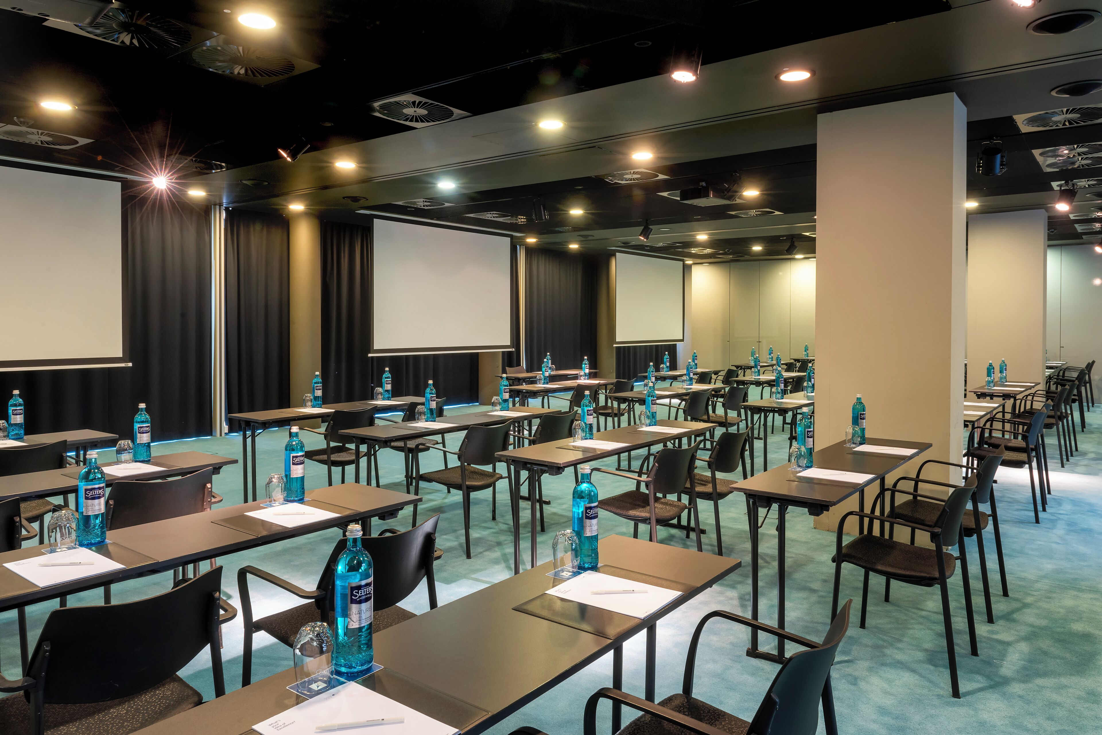 Una sala conferenze con file di tavoli, sedie e schermi di proiezione.
