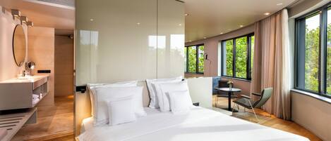 A modern hotel room with a large bed, a desk, and a chair, a bathroom with a mirror and shelves, and large windows with a view of trees.