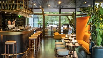 A modern bar area with a bartender, bar stools, and a wine rack. There are seating areas with tables and chairs, and large windows offering a view of the outdoors.