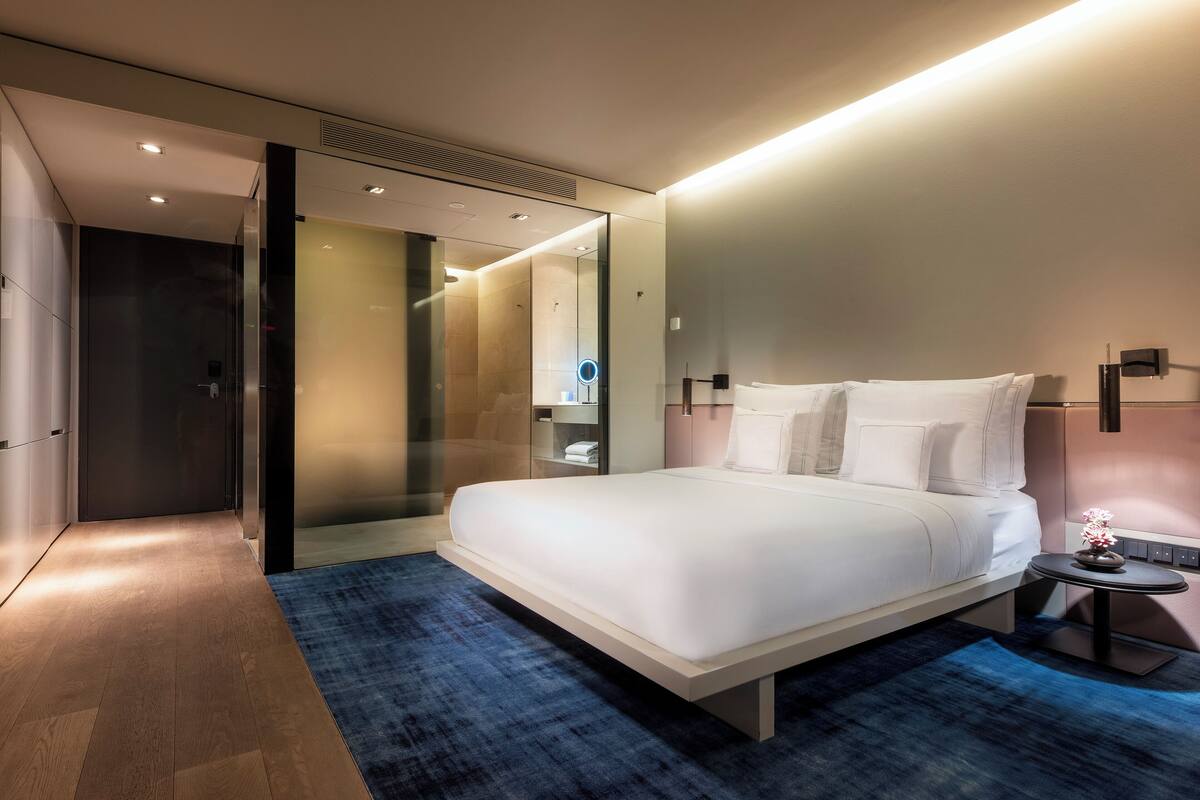 Moderni hotellihuone, jossa on suuri sänky, pieni yöpöytä, peili ja lasiseinällä erotettu suihkutila.