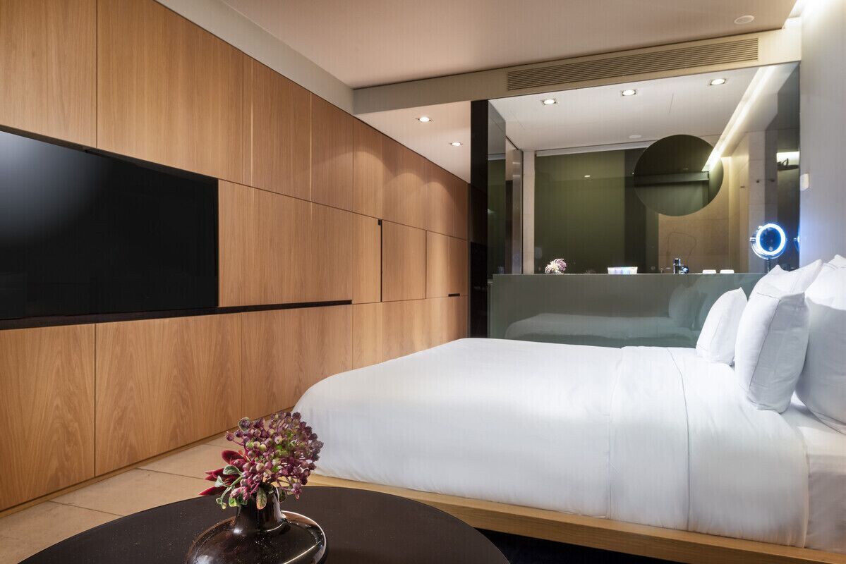 Una habitación de hotel moderna con una cama grande, un panel de pared de madera, una mesa de centro redonda con un jarrón de flores y un espejo grande con luz circular.