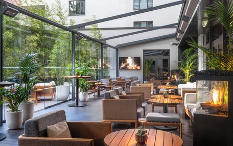 A spacious outdoor seating area with multiple tables and chairs, surrounded by large potted plants and a fire feature.