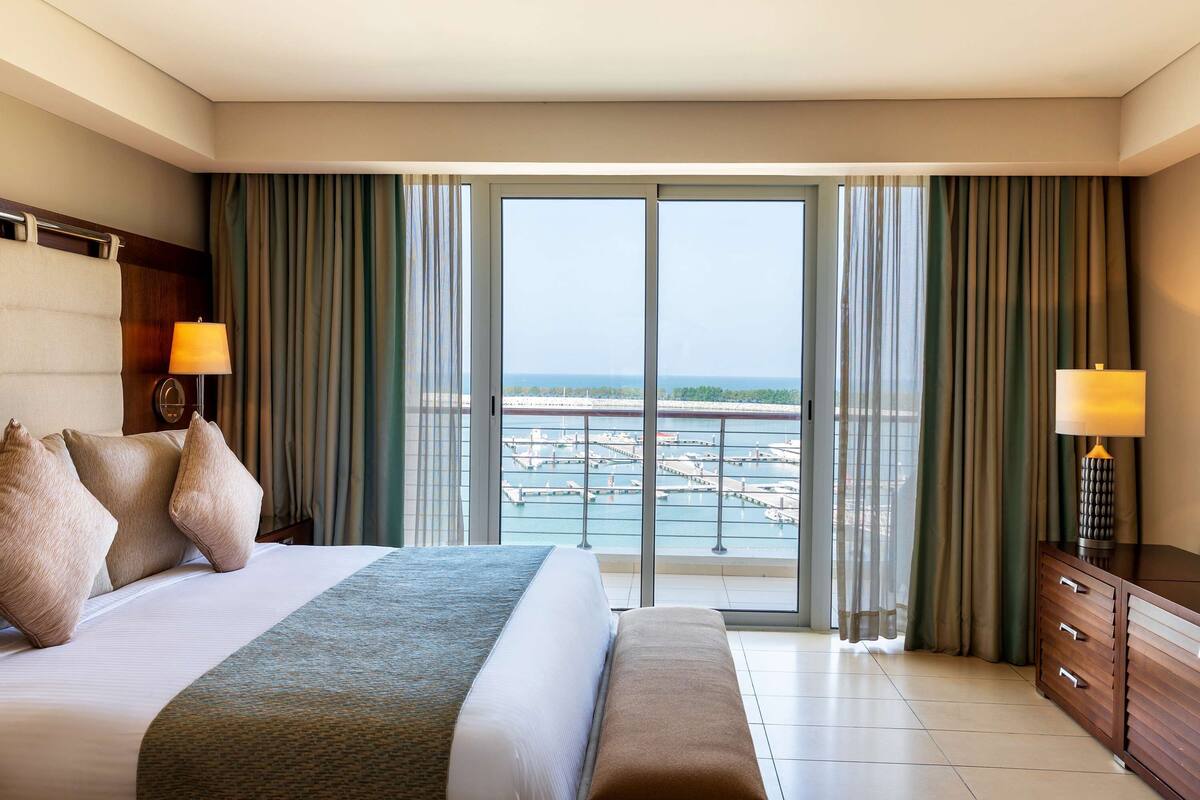 Quarto de hotel com uma cama grande, um banco, uma mesinha de cabeceira com abajur e vista para uma marina através de uma porta de vidro deslizante.