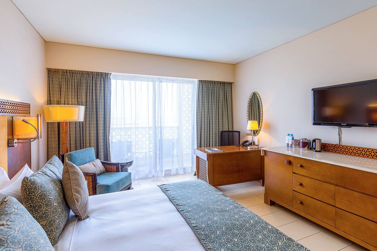 Quarto de hotel com cama, escrivaninha, televisão e janela com cortinas.