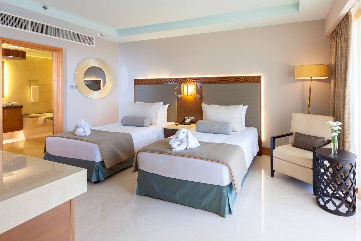 Quarto de hotel com duas camas, um espaço de estar com uma cadeira, um espelho redondo, mesa de cabeceira e abajur.