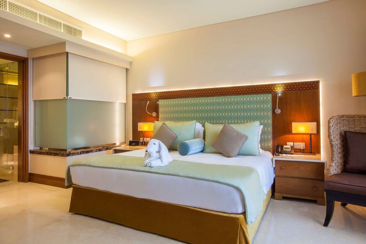 Quarto de hotel com uma cama grande, mesas de cabeceira, uma cadeira e um banheiro com porta de vidro.