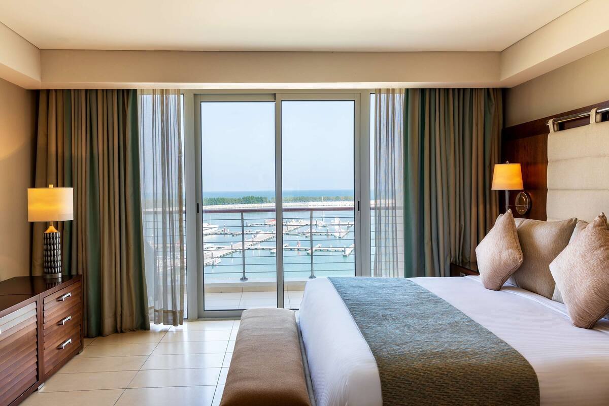 Quarto de hotel com cama grande, um banco, cômoda, duas luminárias e vista para um porto através de uma porta de correr de vidro.
