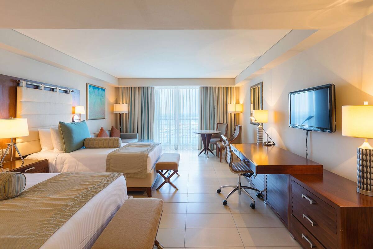 Quarto de hotel com duas camas, uma escrivaninha, uma cadeira, uma televisão e janela com cortinas.