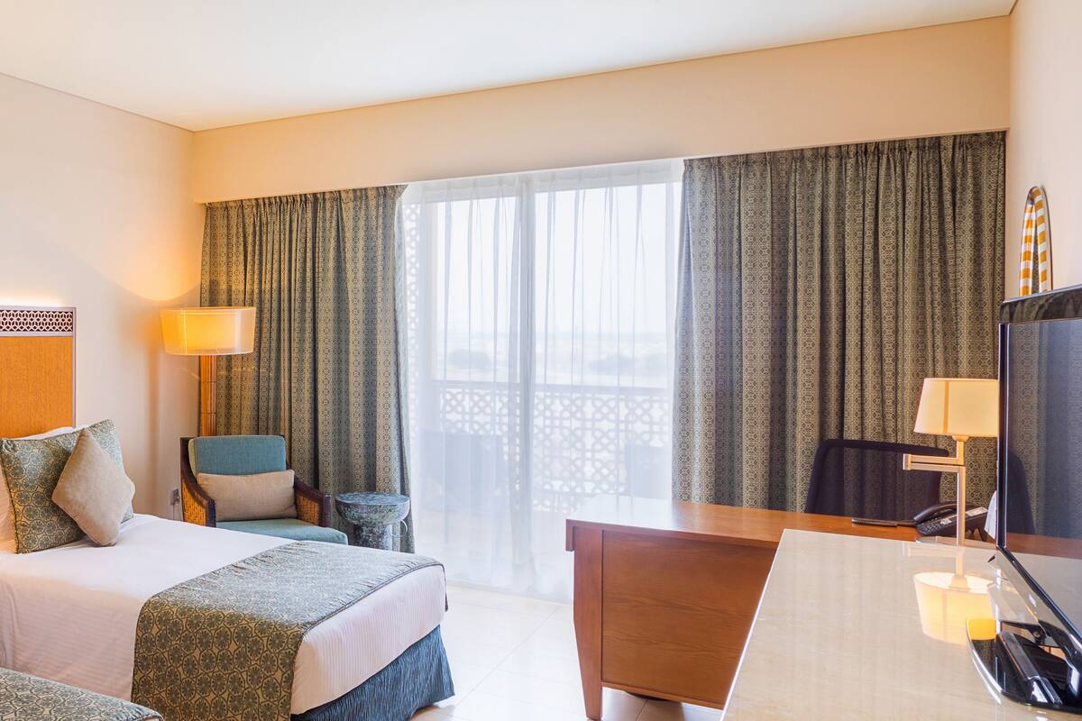 Quarto de hotel com cama, escrivaninha, cadeira, televisão e varanda com cortinas.