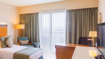 Habitación de hotel con cama, escritorio, silla, televisor y balcón con cortinas.