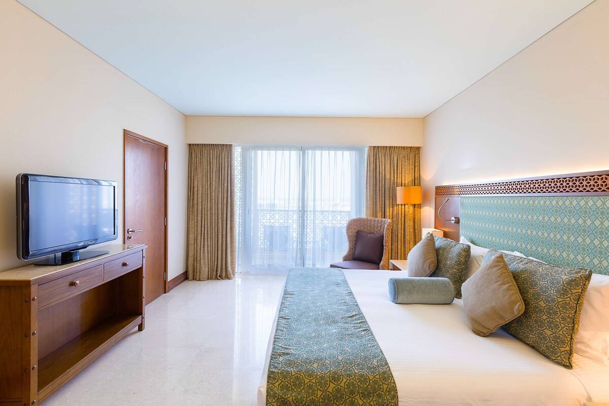 Quarto de hotel com televisão em um armário de madeira, cama com almofadas estampadas, sofá com almofadas, janela com cortinas e cabeceira estampada.