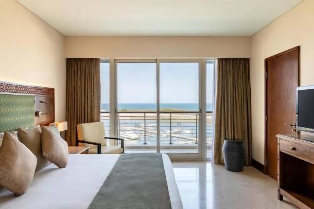 Quarto de hotel com cama, poltrona, televisão, janela grande com vista para a praia e varanda.