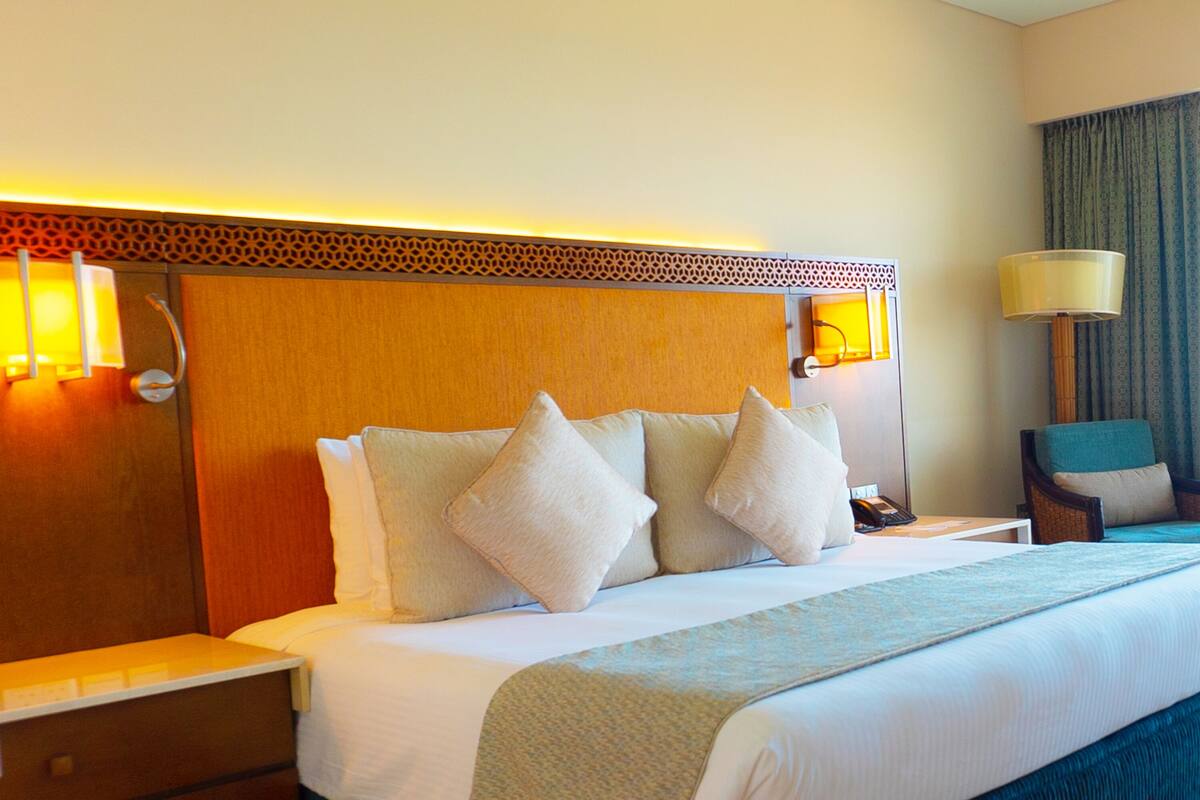 Quarto de hotel com uma cama grande, cabeceira de madeira, luminárias laterais, uma cadeira e uma mesa de cabeceira.