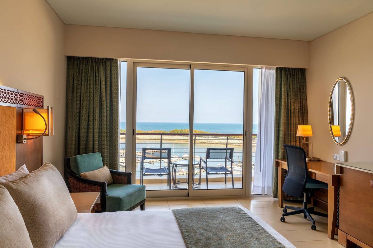 Quarto de hotel com uma cama, uma escrivaninha, uma cadeira e vista para a praia através de uma porta de correr.