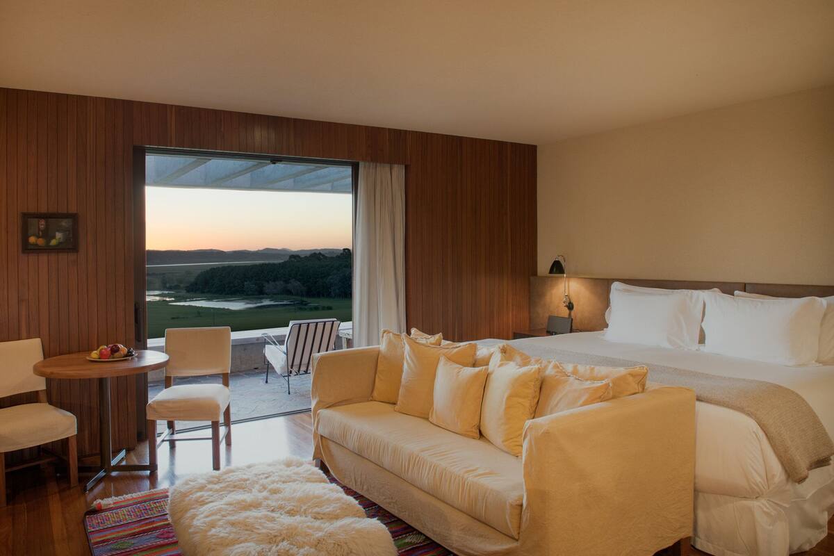 Habitación de hotel con una cama grande, un sofá, una mesita con fruta y vistas al exterior.