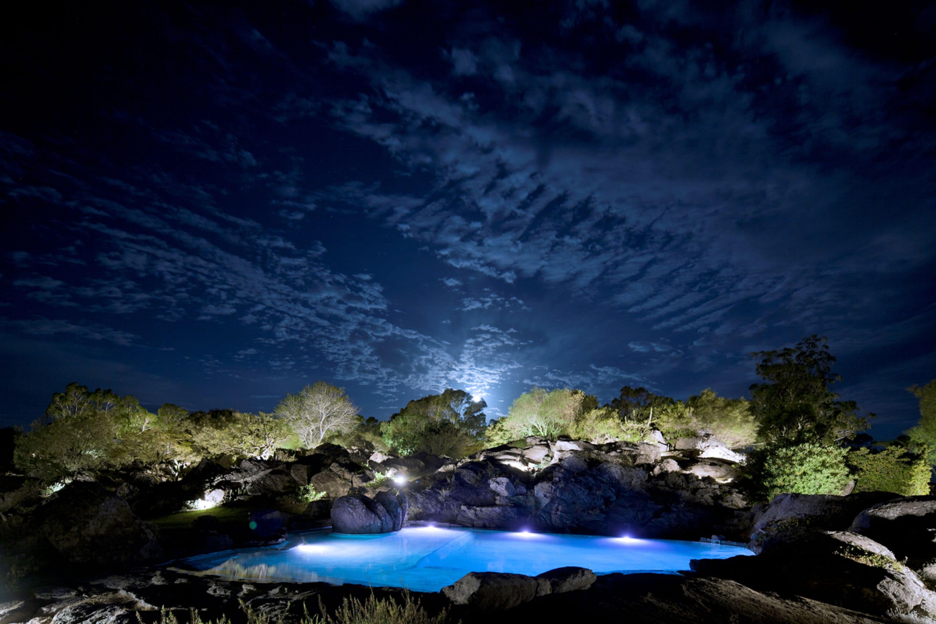 A nighttime pool area with a clear sky, surrounded by rocks and trees, and illuminated by artificial lights.