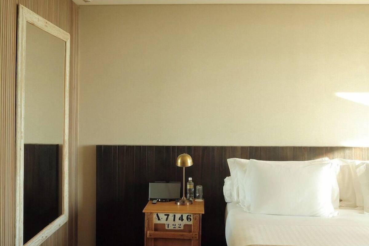 Habitación de hotel con cama, mesita de noche con lámpara, televisor y cabecero de madera.