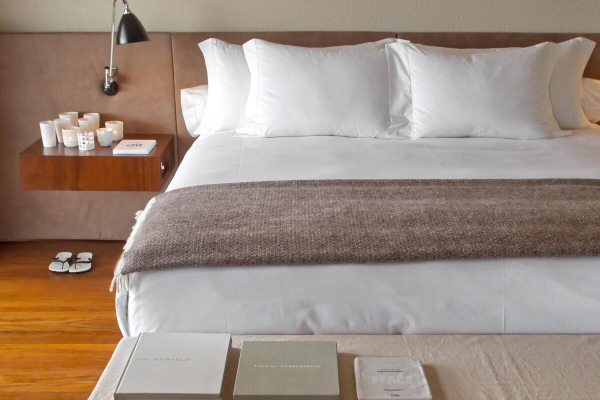 Una cama bien hecha con sábanas blancas, un plaid marrón y una mesita de noche con una lámpara, velas y un libro.
