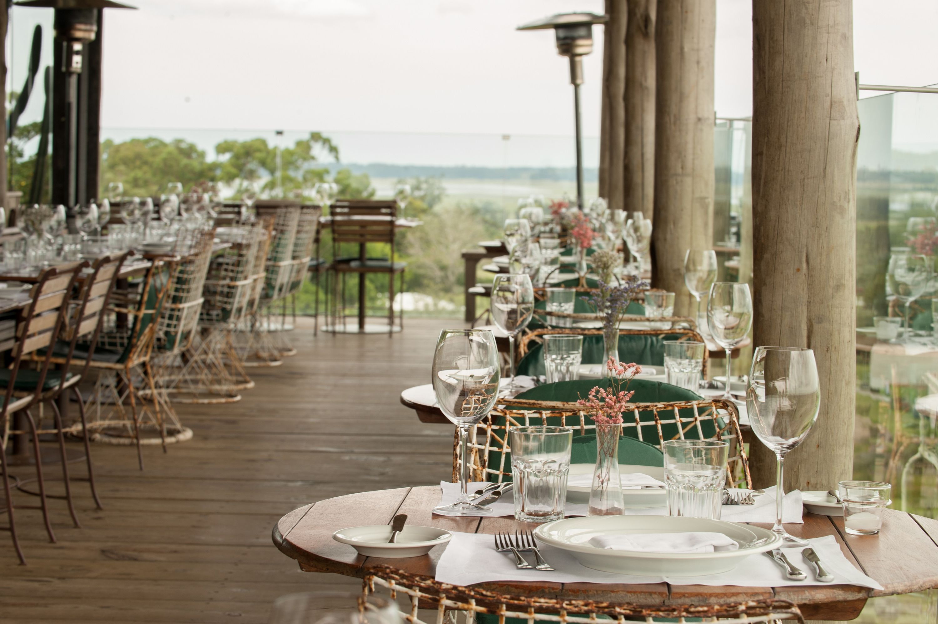 A long dining table set for a meal with chairs on a wooden deck, overlooking a scenic landscape.