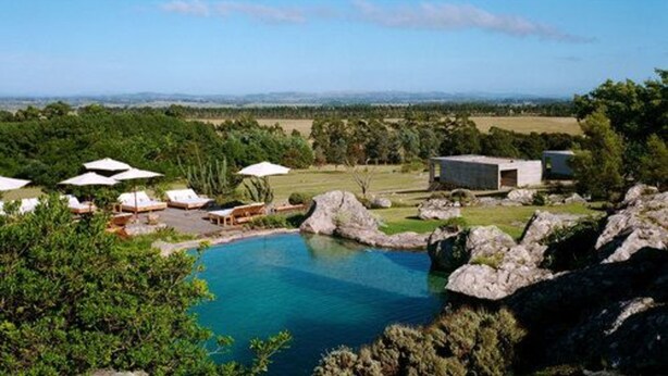Un área de piscina con sillones de descanso y sombrillas, rodeada de rocas y follaje.