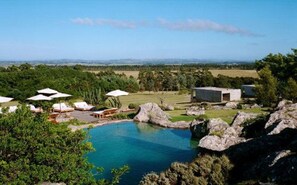 Un área de piscina con sillones de descanso y sombrillas, rodeada de rocas y follaje.