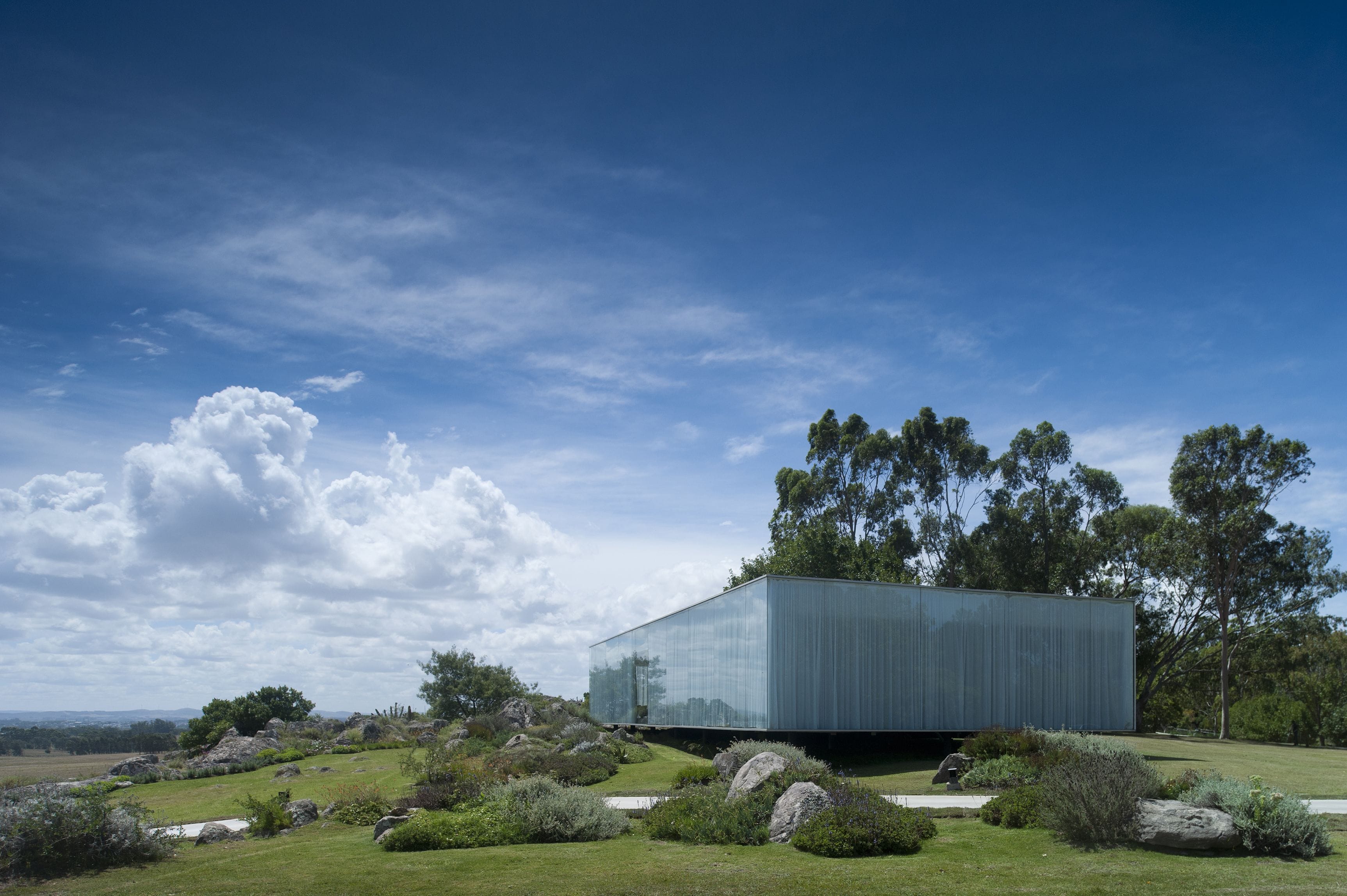 Un bâtiment moderne à la façade en verre réfléchissant, entouré d’un jardin aménagé avec des rochers et des arbustes, sous un ciel bleu dégagé.