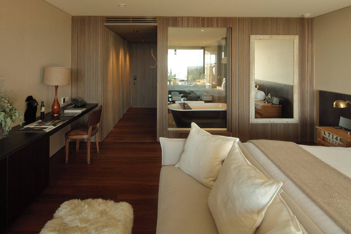 Una habitación de hotel con cama, escritorio, silla, alfombra pequeña y espejo.