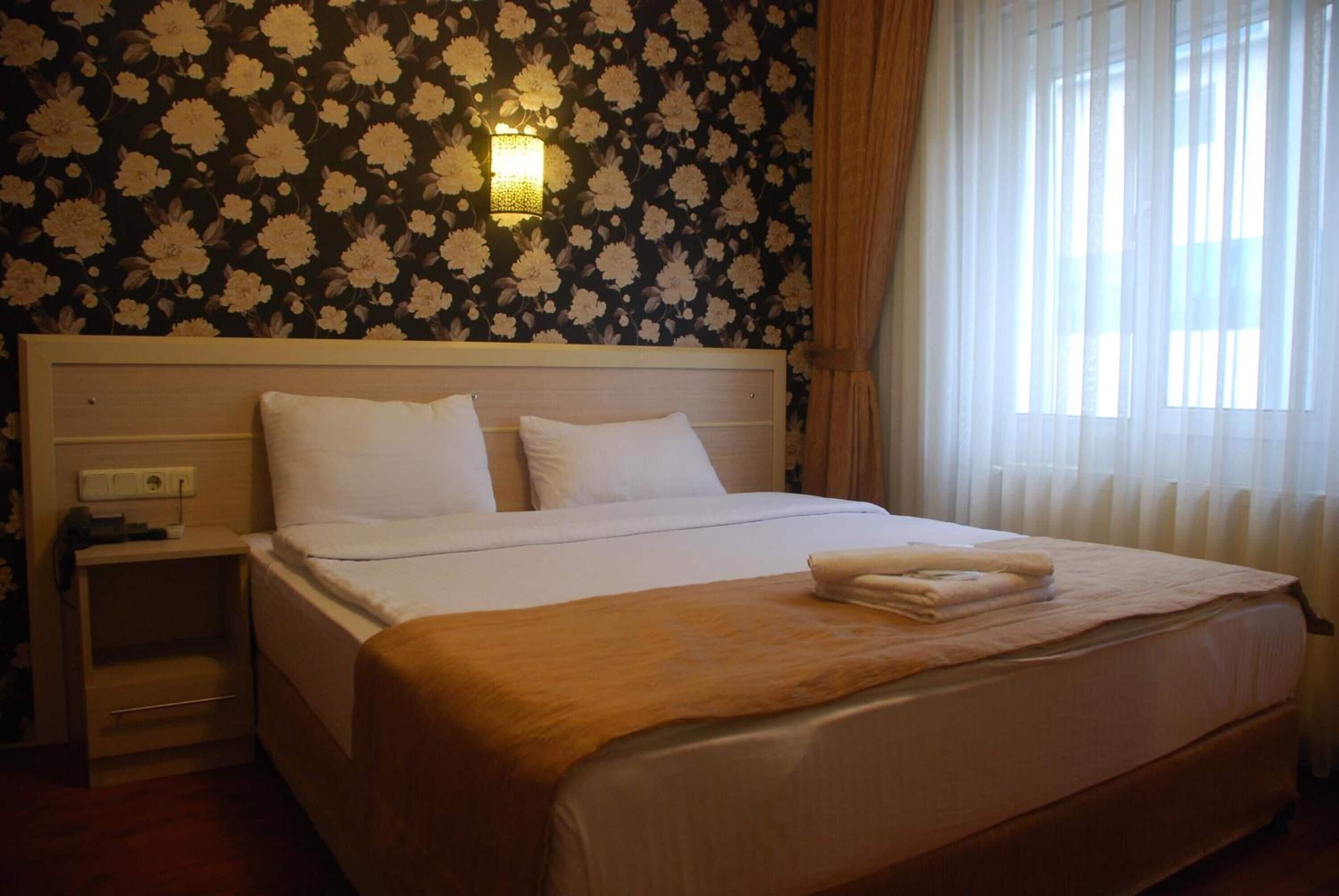 A hotel room with a large bed, a nightstand, a wall-mounted light, and a window with curtains.