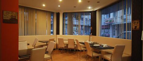 Un comedor con mesas y sillas, ventanas amplias con persianas, y vista a la calle.
