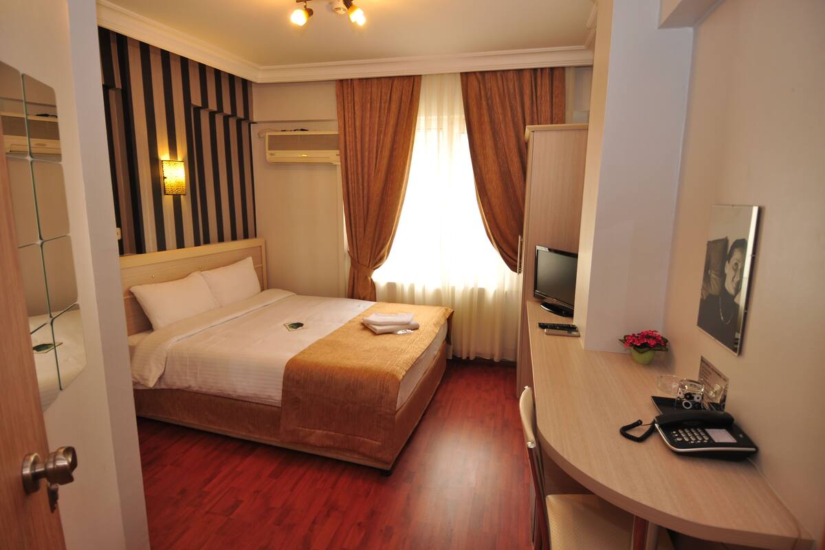 A hotel room with a bed, a desk with a phone, a TV, and a window with curtains.